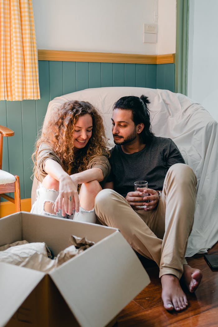 Cheerful woman in trendy jeans speaking with content ethnic barefoot boyfriend with glass of water while sitting on comfortable couch near open cardboard box with parchment in flat